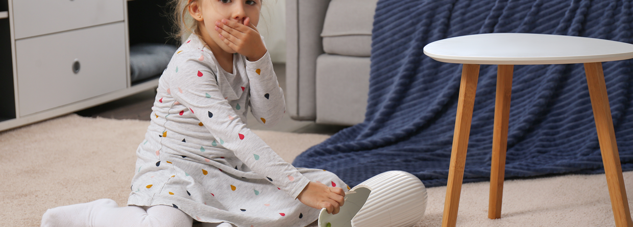 Een klein meisje schrikt en heeft haar hand voor haar mond omdat de vaas met bloemen gevallen en gebroken is